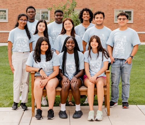 Teens pose for a photo while sitting on an outdoor bench, wearing blue MBV t-shirts and smiling.