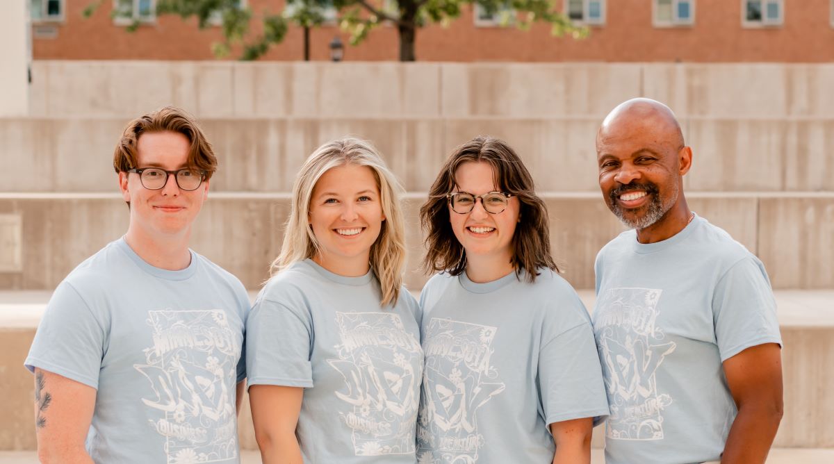 Four adults pose for a photo together, wearing blue MBV-shirts and smiling.