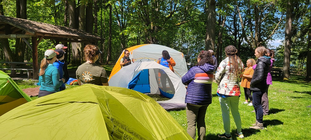 Group of educators stand in a cricle while the facilitator demonstrates how to set up a tent