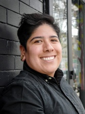 Representative Samantha Montaño's headshot, standing and smiling in front of a black brick wall.